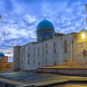 The blue dome and brown mud structures of the Kalon Mosque and minaret under cloudy, dusk skies.