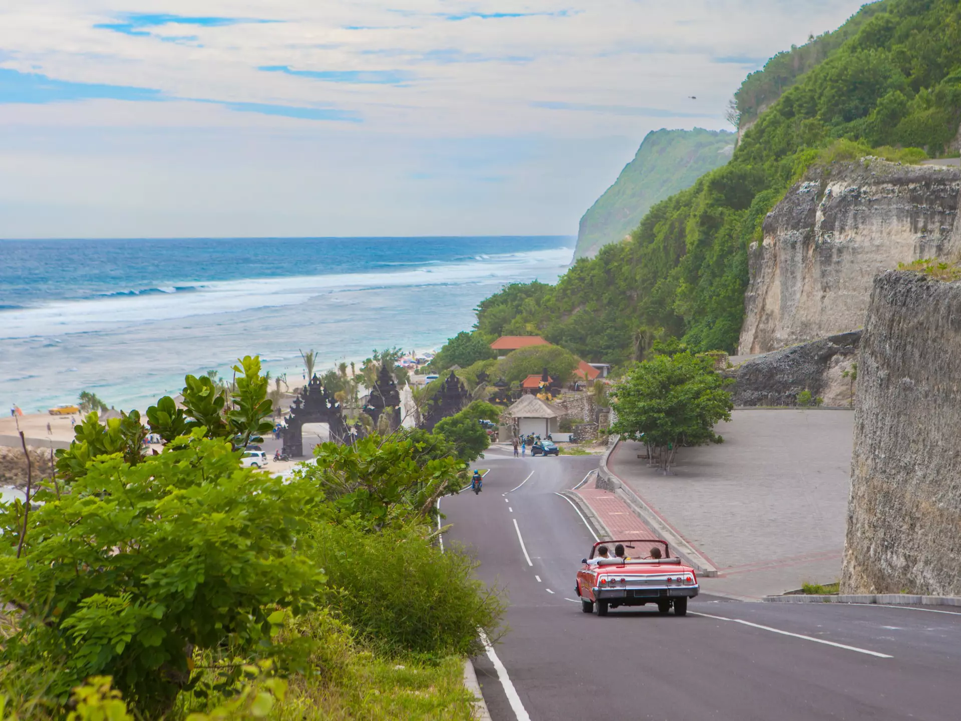 A red car drives down a hill towards a beach in a tropical destination