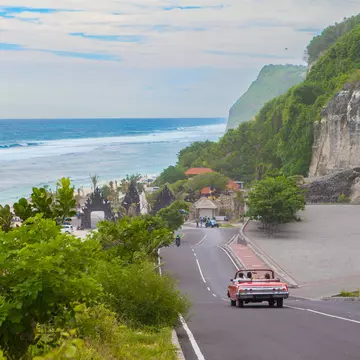 A red car drives down a hill towards a beach in a tropical destination