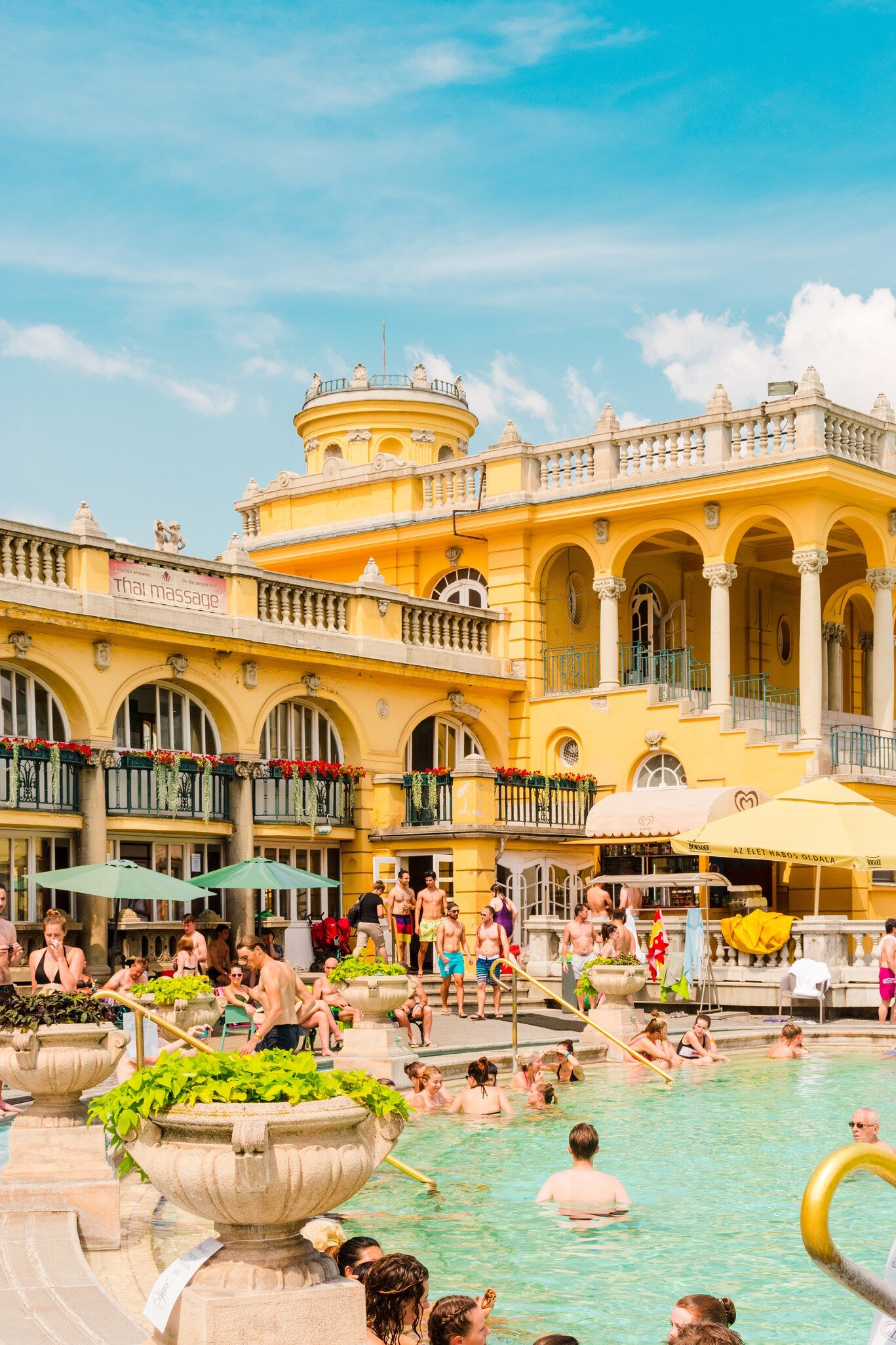 People relax in an outdoor pool overlooked by an ornate yellow building.