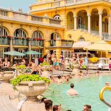 People relax in an outdoor pool overlooked by an ornate yellow building.