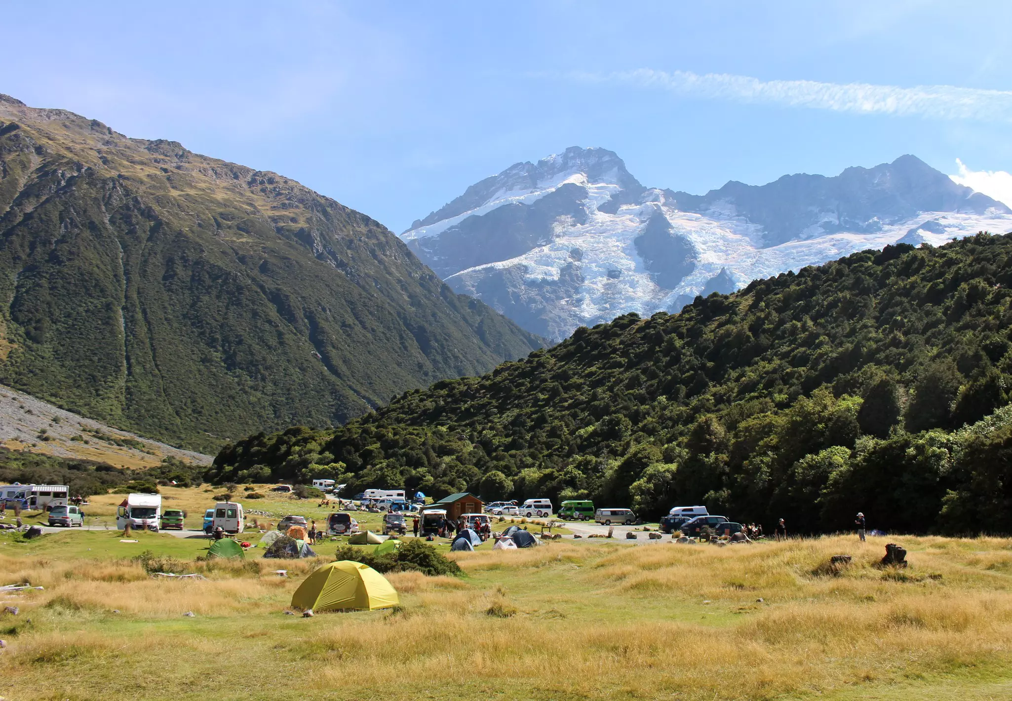 Tents pitched in grass and vans and cars parked on pavement in a mountainous part of New Zealand.