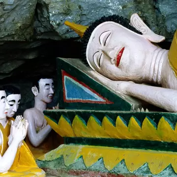 A painted plaster reclining Buddha on a green and yellow plinth in a cave, three small kneeling figures with hands pressed together before it.