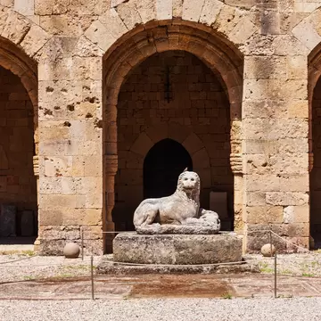 Weathered marble lion on a plinth before three stone arches, with a roped-off mosaic floor and gravel in front