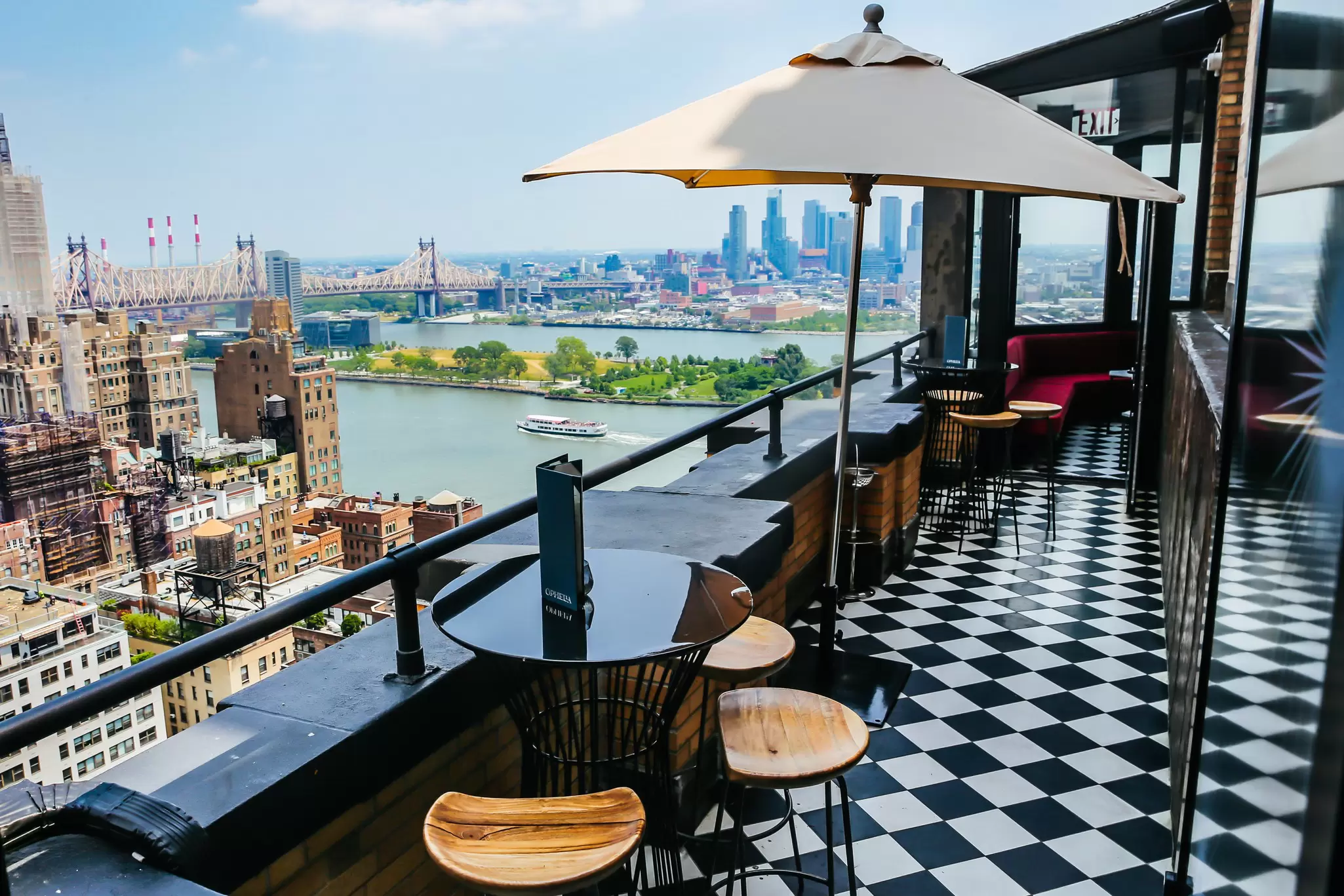 Small round tables with chairs are on a roofdeck with a black-and-white checked floor; a river with a boat and tall buildings on the other shore are in the background.