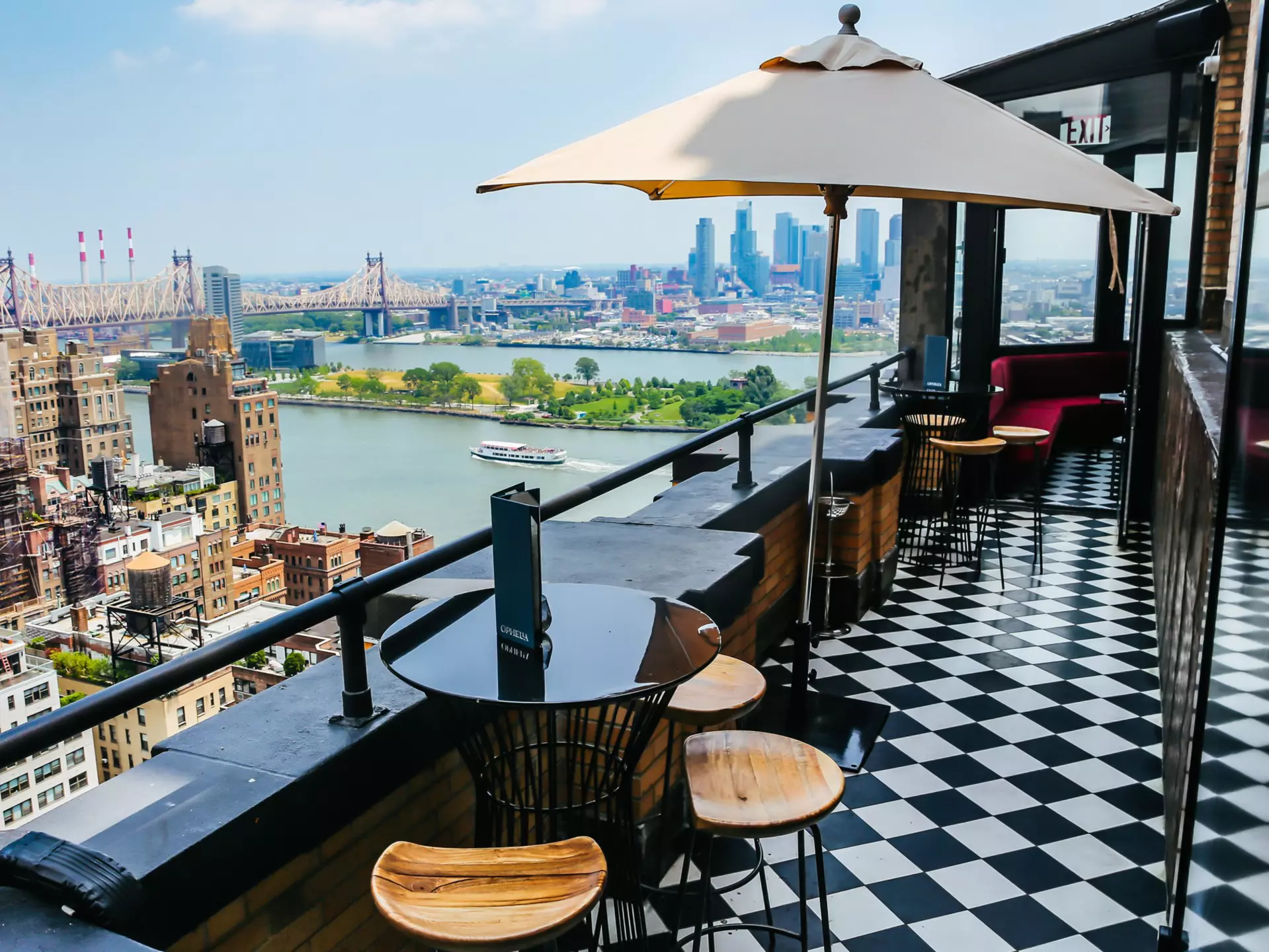 Small round tables with chairs are on a roofdeck with a black-and-white checked floor; a river with a boat and tall buildings on the other shore are in the background.