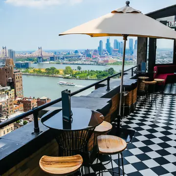 Small round tables with chairs are on a roofdeck with a black-and-white checked floor; a river with a boat and tall buildings on the other shore are in the background.