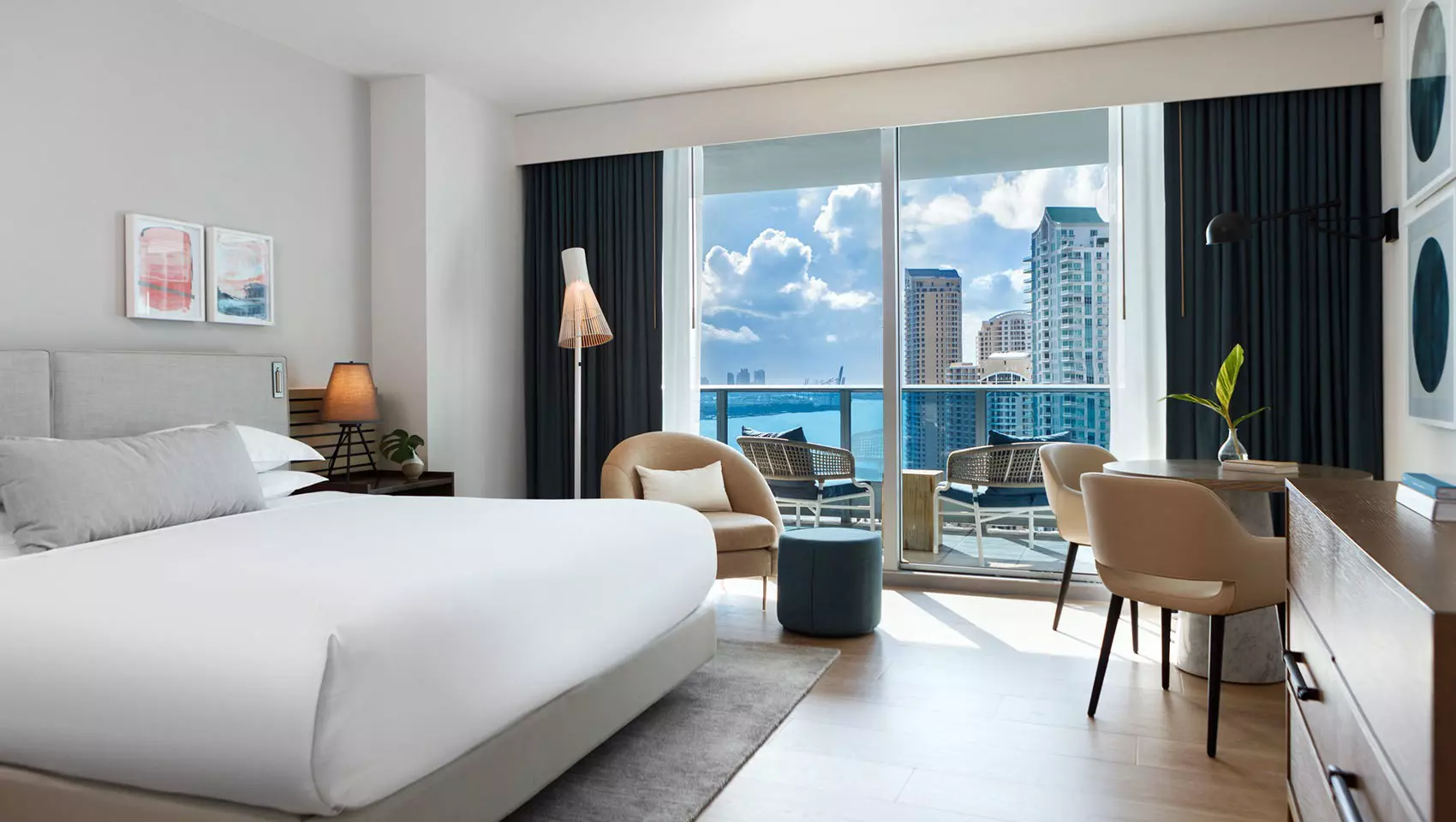 A room at the Kimpton Epic hotel in Miami has a curved class wall made up of five narrow rectangular panes overlooking a sea of skyscrapers outside. Inside, a bed faces the curved window wall, made up with simple white linens on a grey woven rug. A tv is mounted to the ceiling and advertises wellness packages. A lamp and chair are just visible in the foreground.