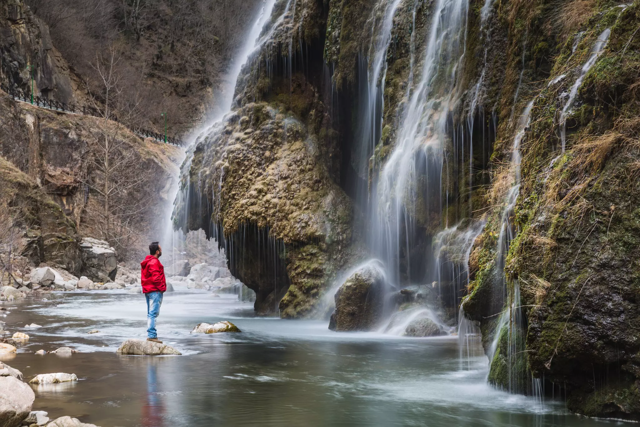 A man in red wet-weather gear and hiking boots stands on a rock in a pool watching a waterfall
