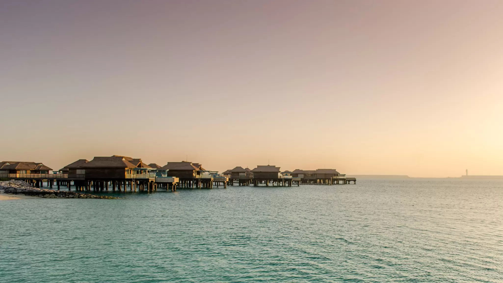 A row of thatched overwater villas stretches across calm sea under a pink and orange sunset sky.