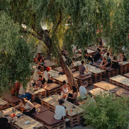 People dine at wooden tables under a canopy of trees.