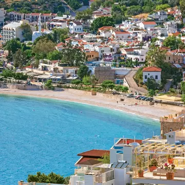 Hillside town of white houses with red roofs and green trees above a curving beach and bright turquoise water.