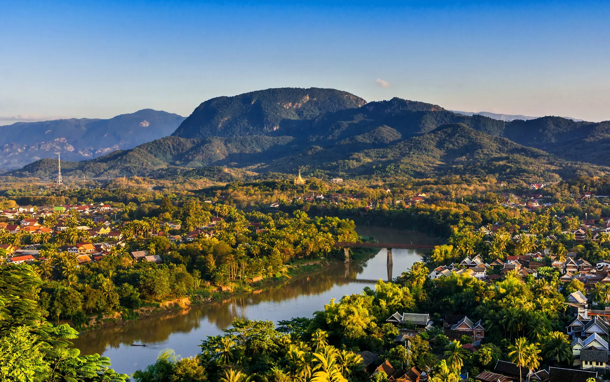 Sunset over a riverside town in Laos set in dense foliage; a range of hills rises in the background.