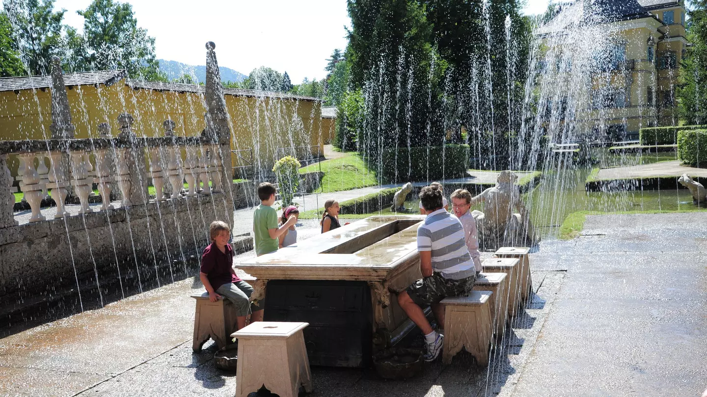 A family sit on ornate stone chairs around a table outdoors in Hellbrunn Palace Park as surprise fountains go off around them