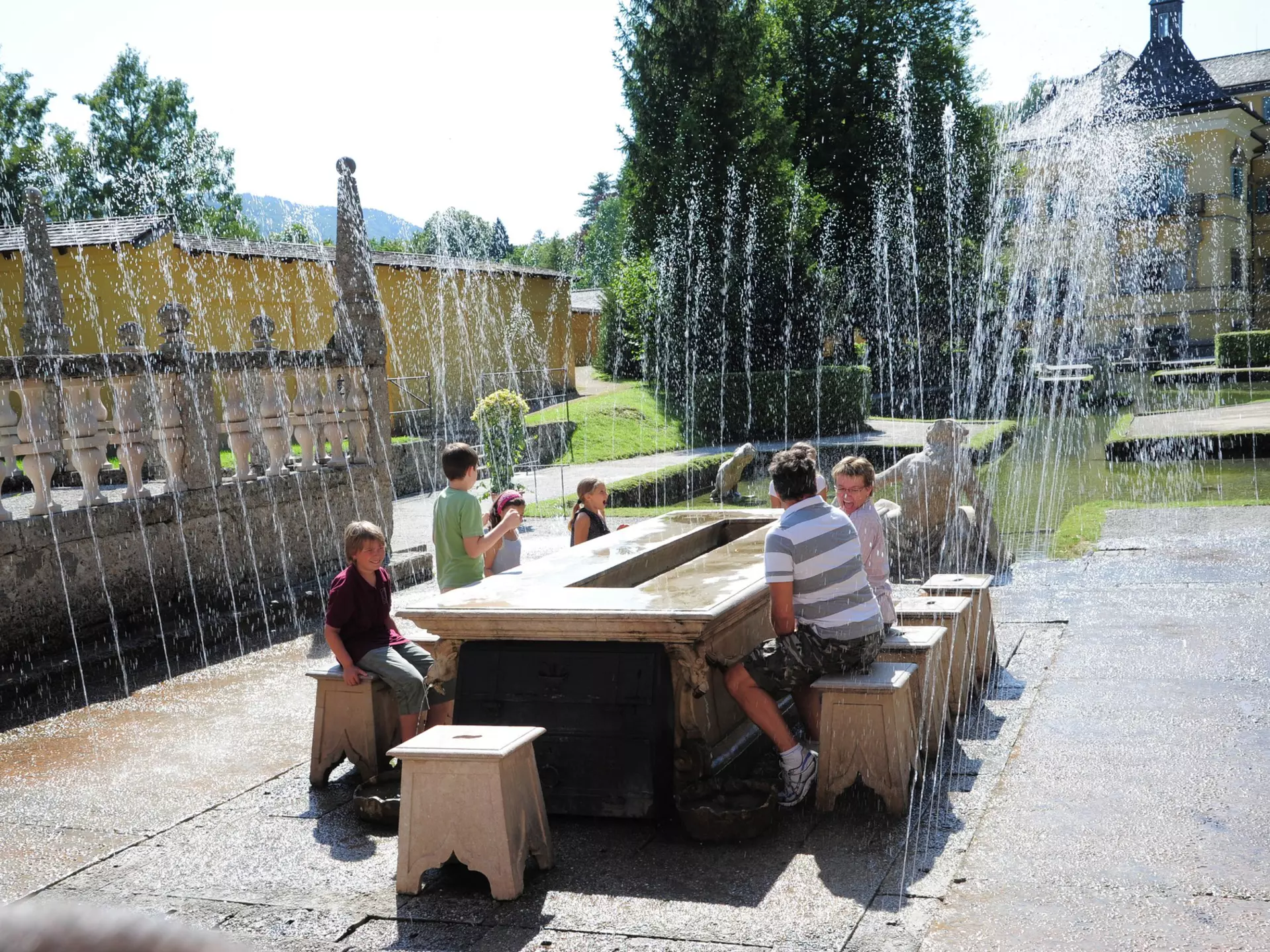 A family sit on ornate stone chairs around a table outdoors in Hellbrunn Palace Park as surprise fountains go off around them