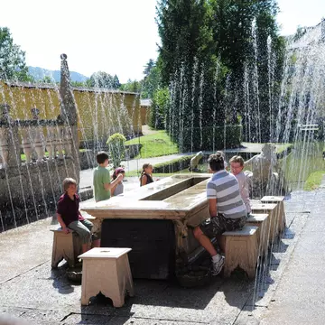 A family sit on ornate stone chairs around a table outdoors in Hellbrunn Palace Park as surprise fountains go off around them