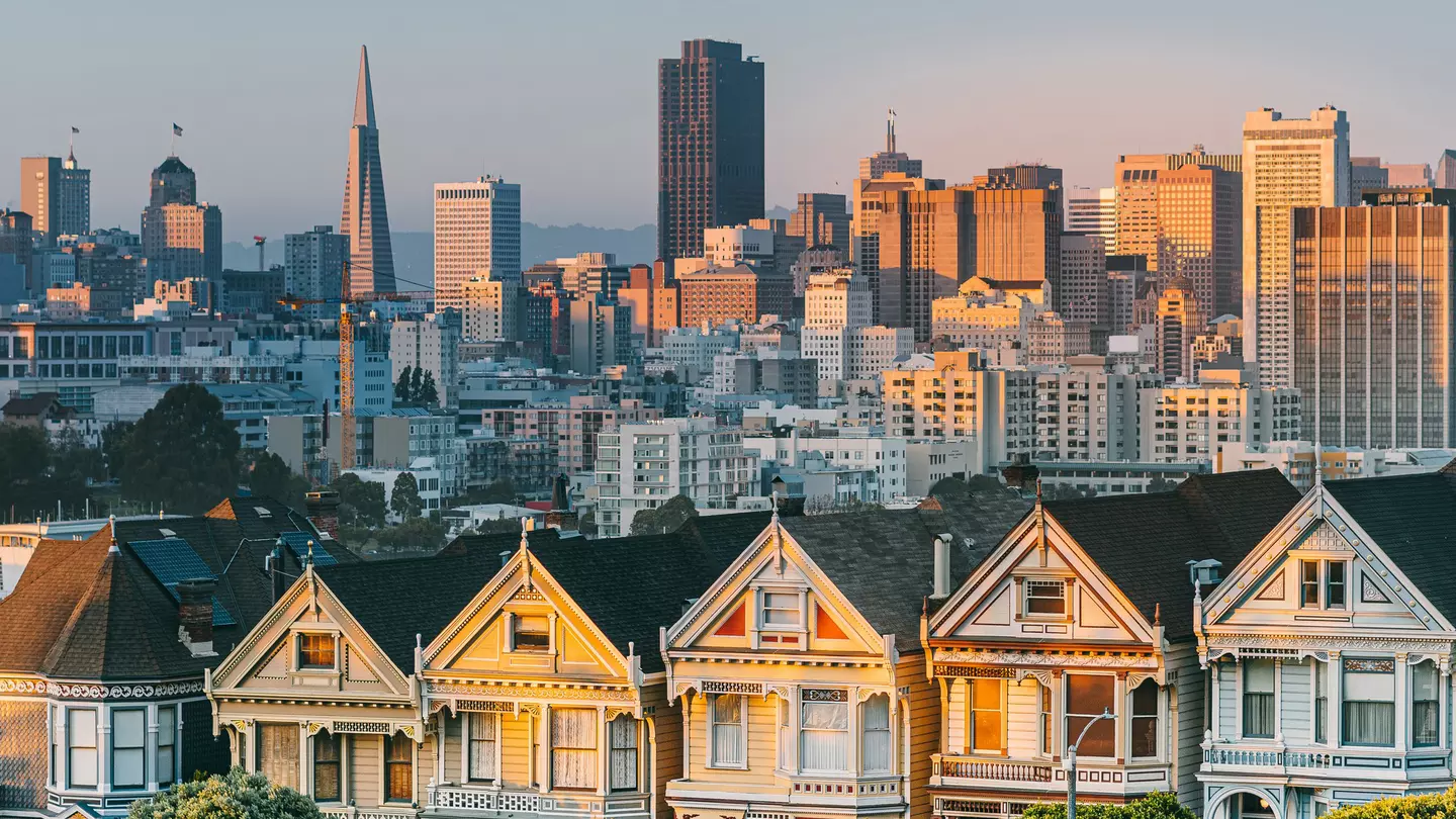 The setting sun shines on painted Edwardian and Victorian homes in a row with the San Francisco city skyline in the distance.