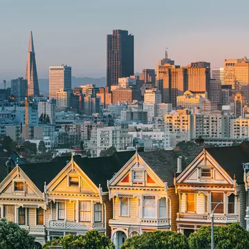 The setting sun shines on painted Edwardian and Victorian homes in a row with the San Francisco city skyline in the distance.