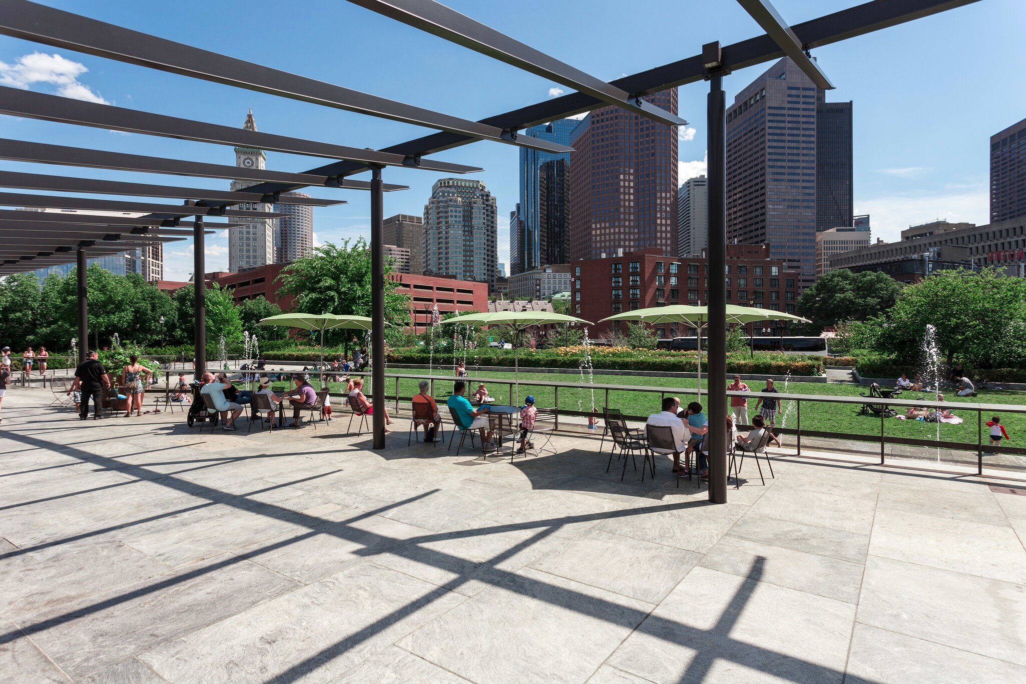 People sitting at tables under umbrellas at the Rose Kennedy Greenway's North End Park in Boston, Massachusetts.
