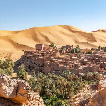 Taghit town ksar of Bechar, Algeria Sahara desert. Palm trees oasis, old dry rocks buildings and sand dune with far tiny tourists and a blue clear sky. From Djebel Baroun rocks in foreground.