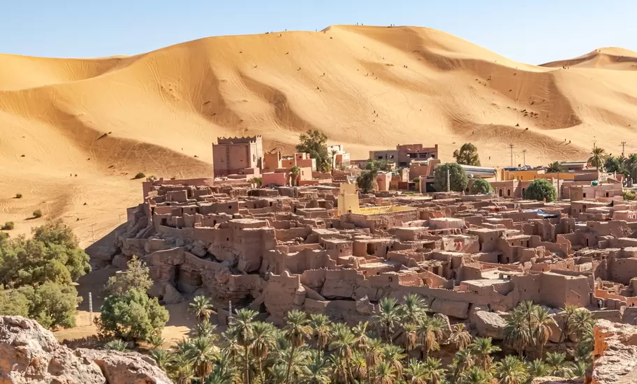 Taghit town ksar of Bechar, Algeria Sahara desert. Palm trees oasis, old dry rocks buildings and sand dune with far tiny tourists and a blue clear sky. From Djebel Baroun rocks in foreground.