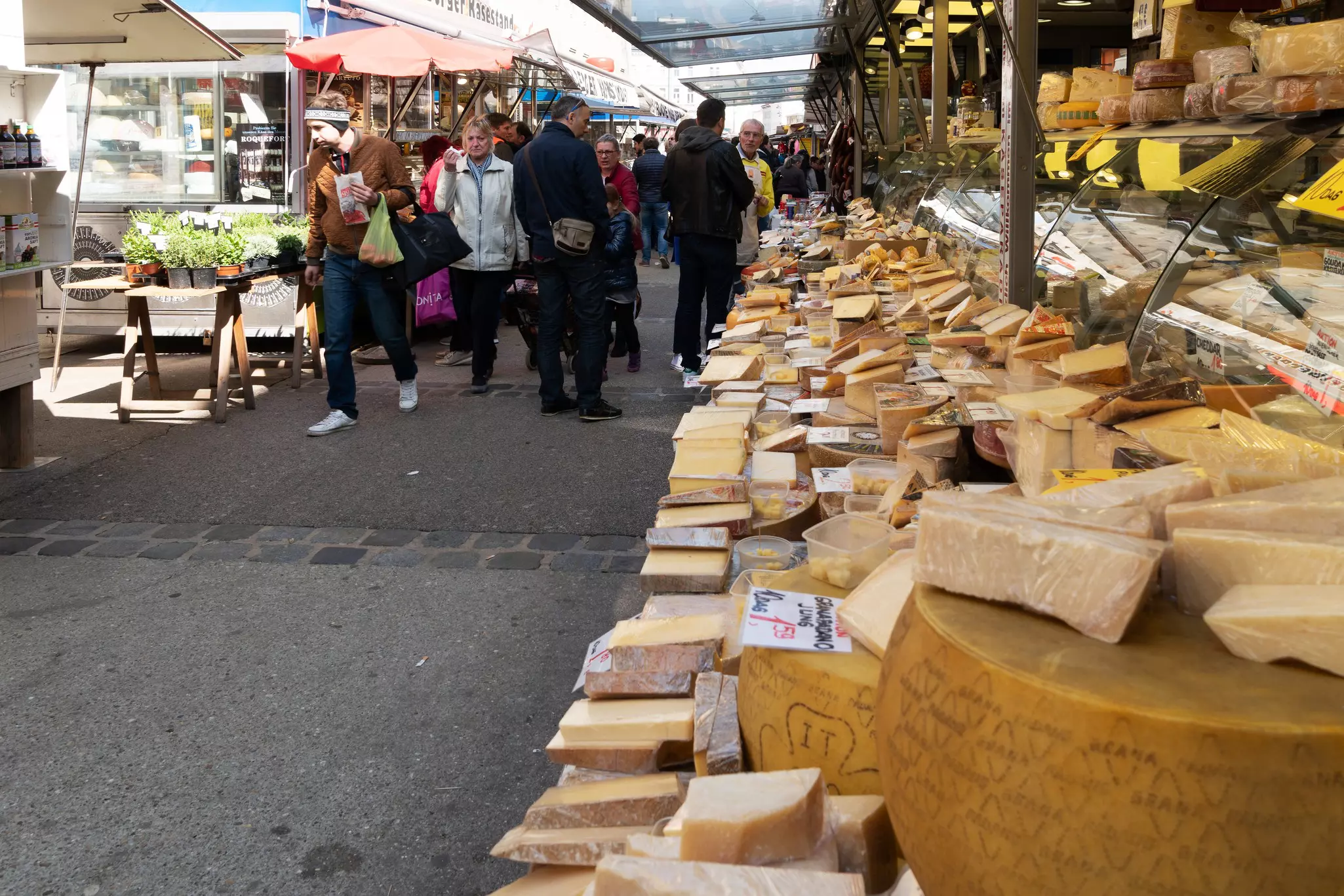 A large display of cheeses is seen at a stall at an outdoor market in a city.
