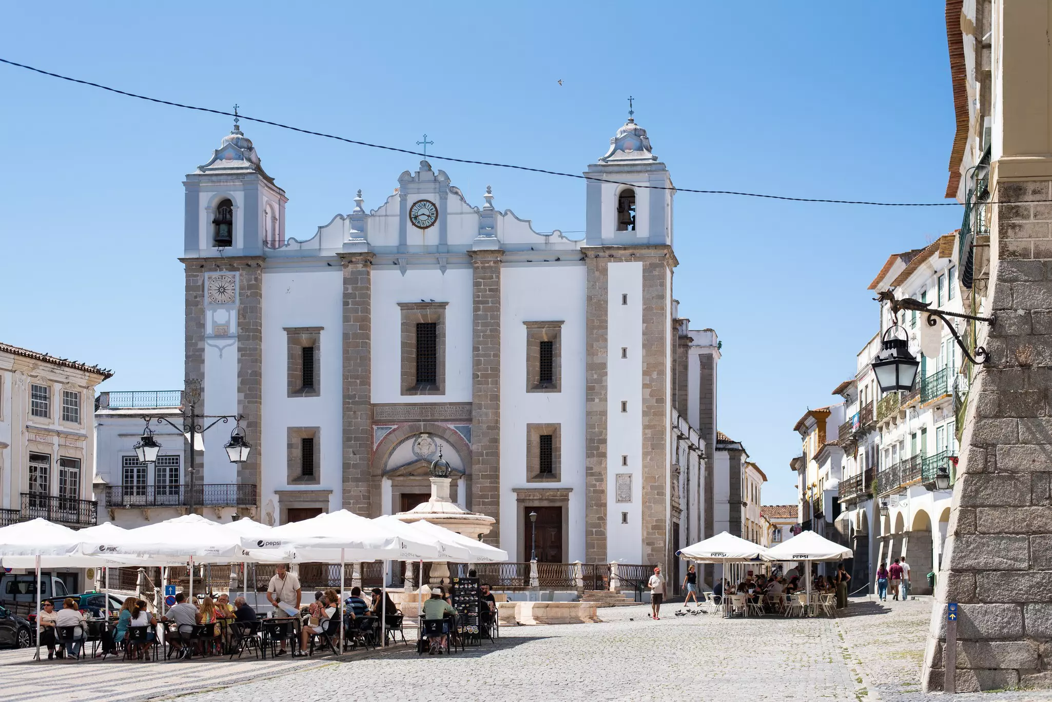 People sit under umbrellas with white-and-stone buildings in the background on a square in Portugal. 