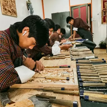 Young men in checked gho bend over carved wooden panels at a long bench laid out with rows of chisels