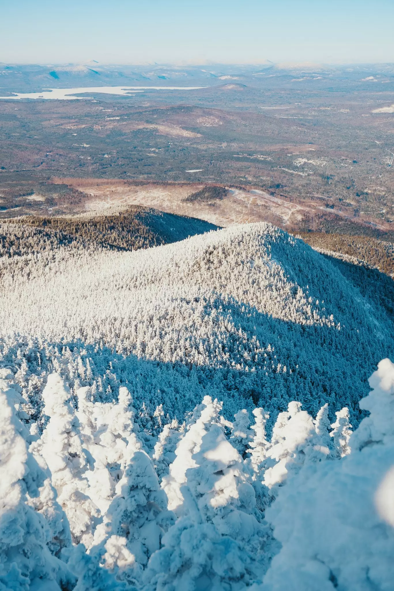 From the top of a mountain in Maine, snow-covered evergreen trees extend down to a valley with no snow.