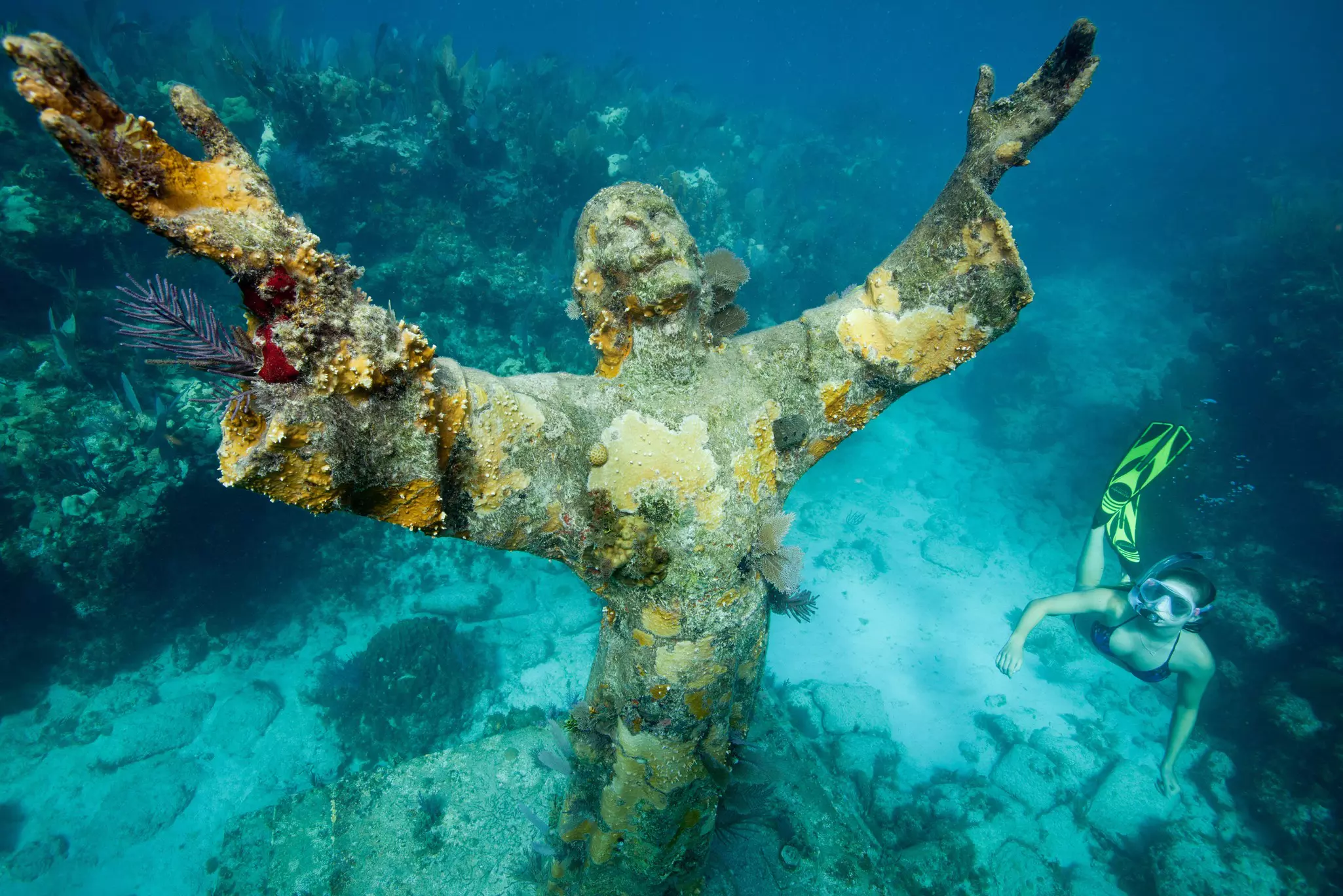 A statue of Jesus Christ sits underwater, encrusted with corals and other marine plants. A snorkeler swims to the right of the statue.