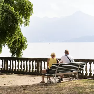 Couple on a green bench beside a stone balustrade on Stresa's lakefront, facing hazy Lake Maggiore and mountains