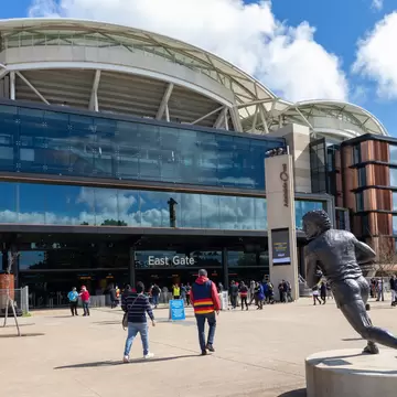 Supporters in club colours walking towards a stadium’s glass entrance under a curved white roof, with a bronze statue of a running footballer in the foreground.