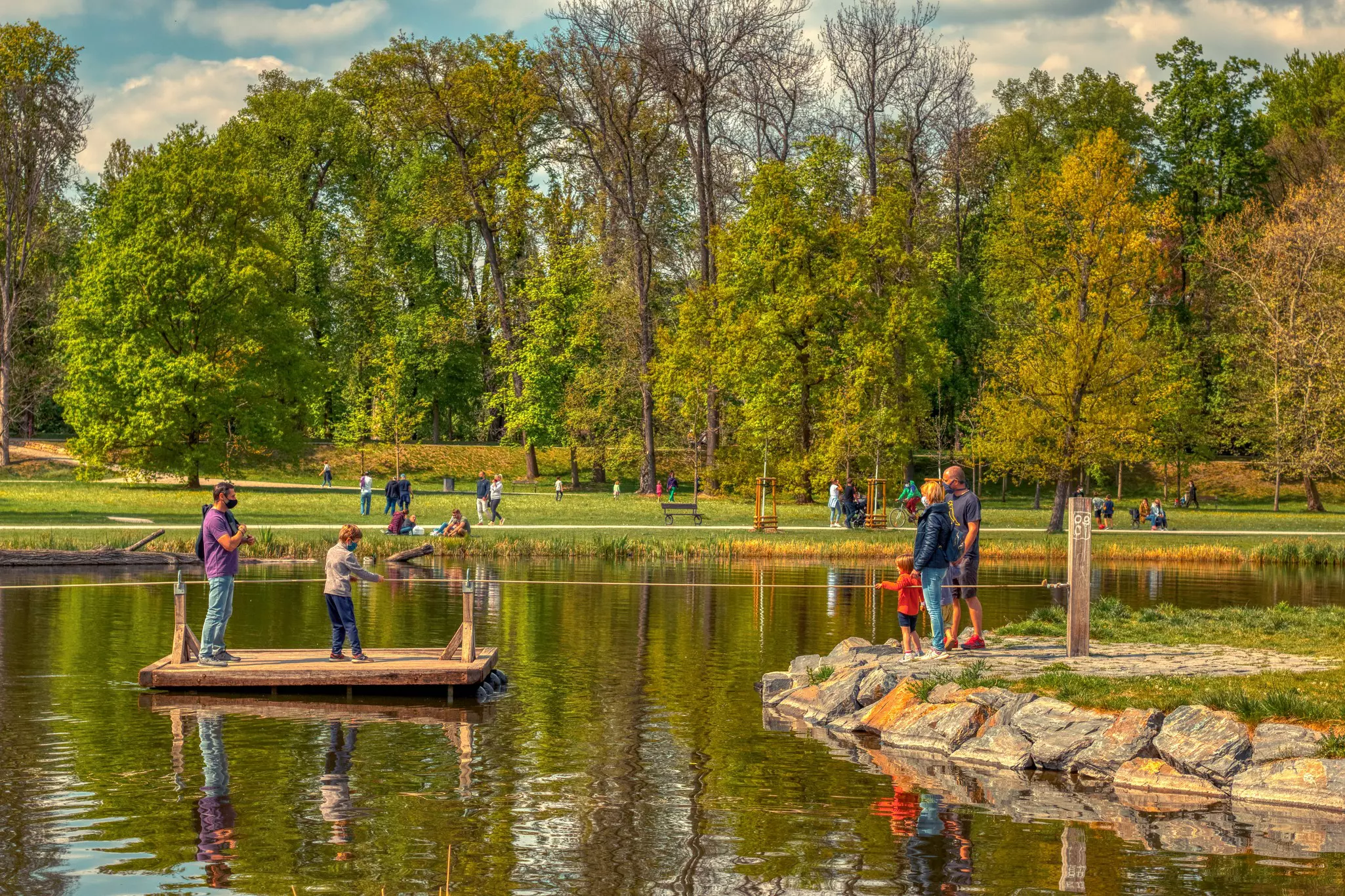 Two people on a wooden raft in a pond pull on a string; three people stand on the shore.