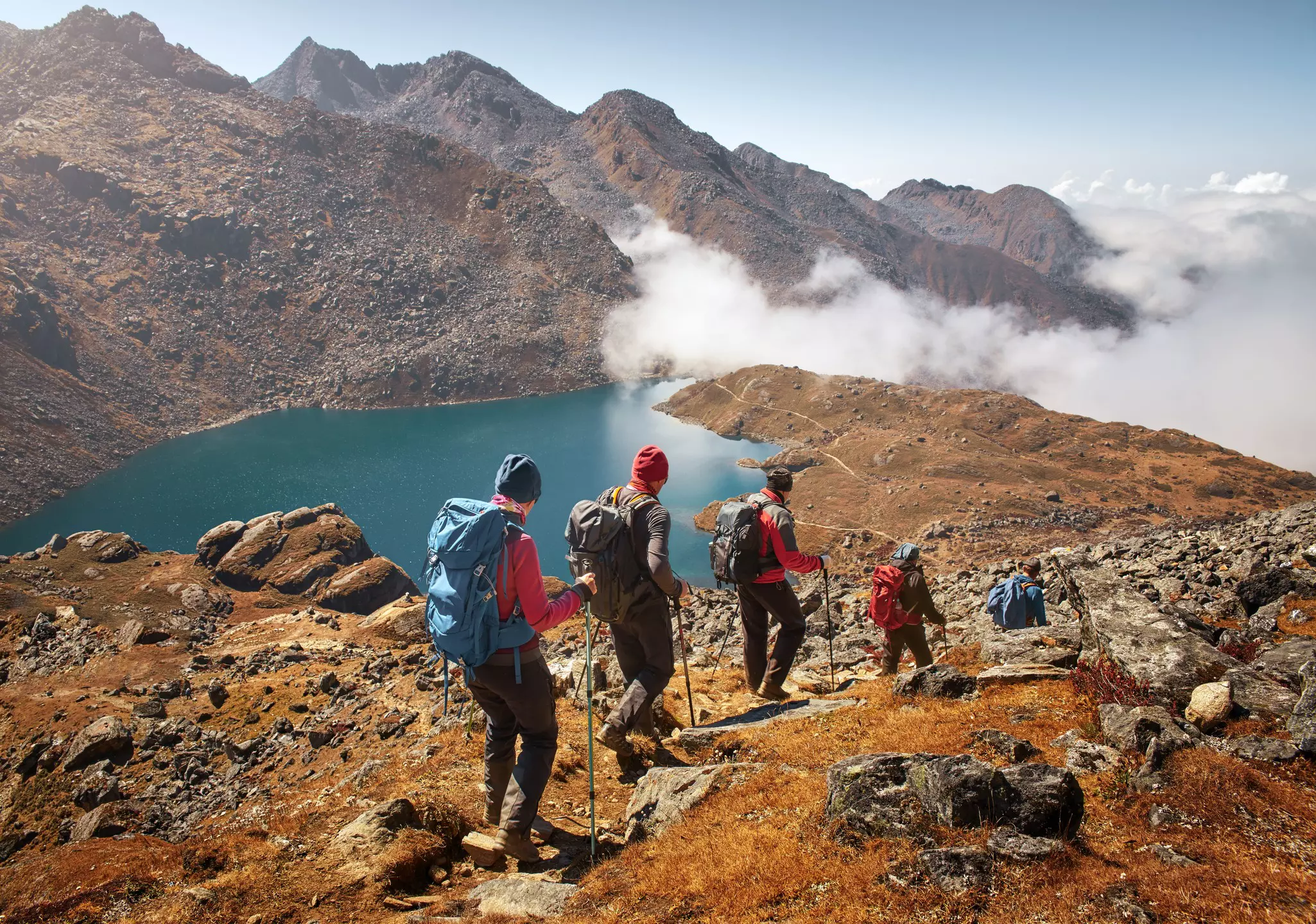 Group of people with backpacks and hiking poles descends down mountain trail to a lake.
