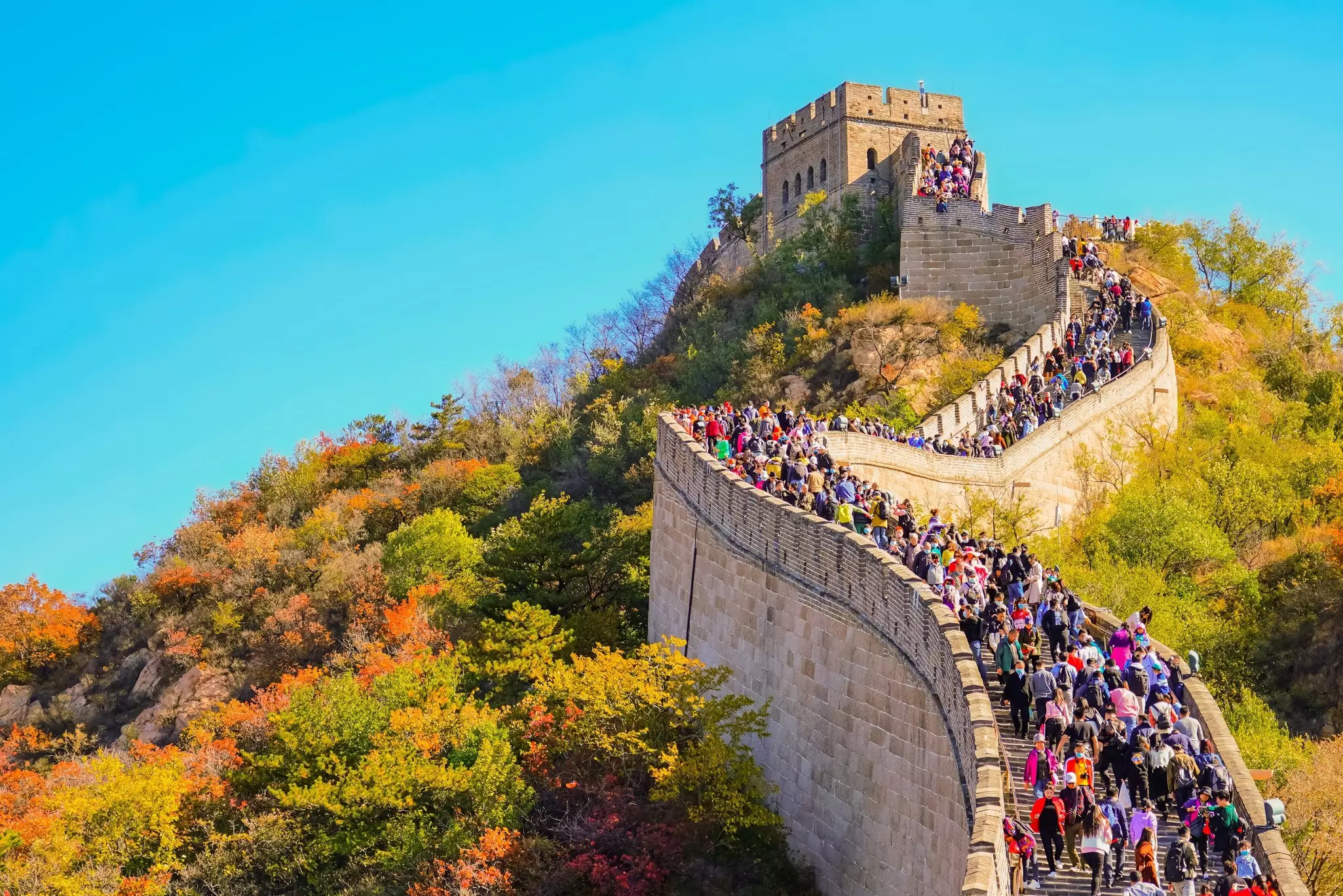 A huge wall snakes up a hill towards a watch tower. Many people are walking on the top of the wall.