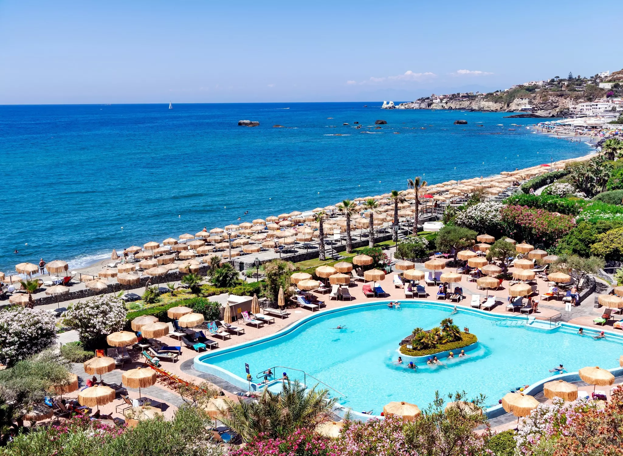 A white-sided oblong pool with light blue water surrounded by umbrellas and lounge chairs, with a beach and the ocean in the distance.