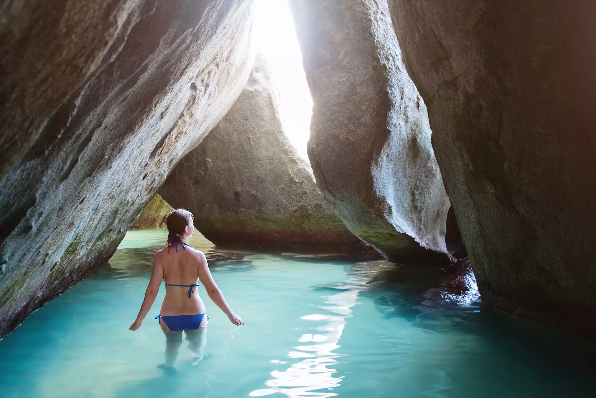 A woman wades in water in a cavern formed by boulders. Sunlight streams in through an opening in the large rocks.