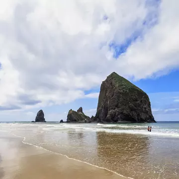 A large freestanding rock at the edge of the ocean. Two people paddle nearby.