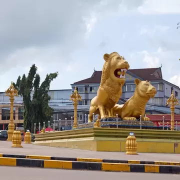 Two golden lion statues on a raised plinth in a traffic circle, ringed by gold lamp posts under grey clouds