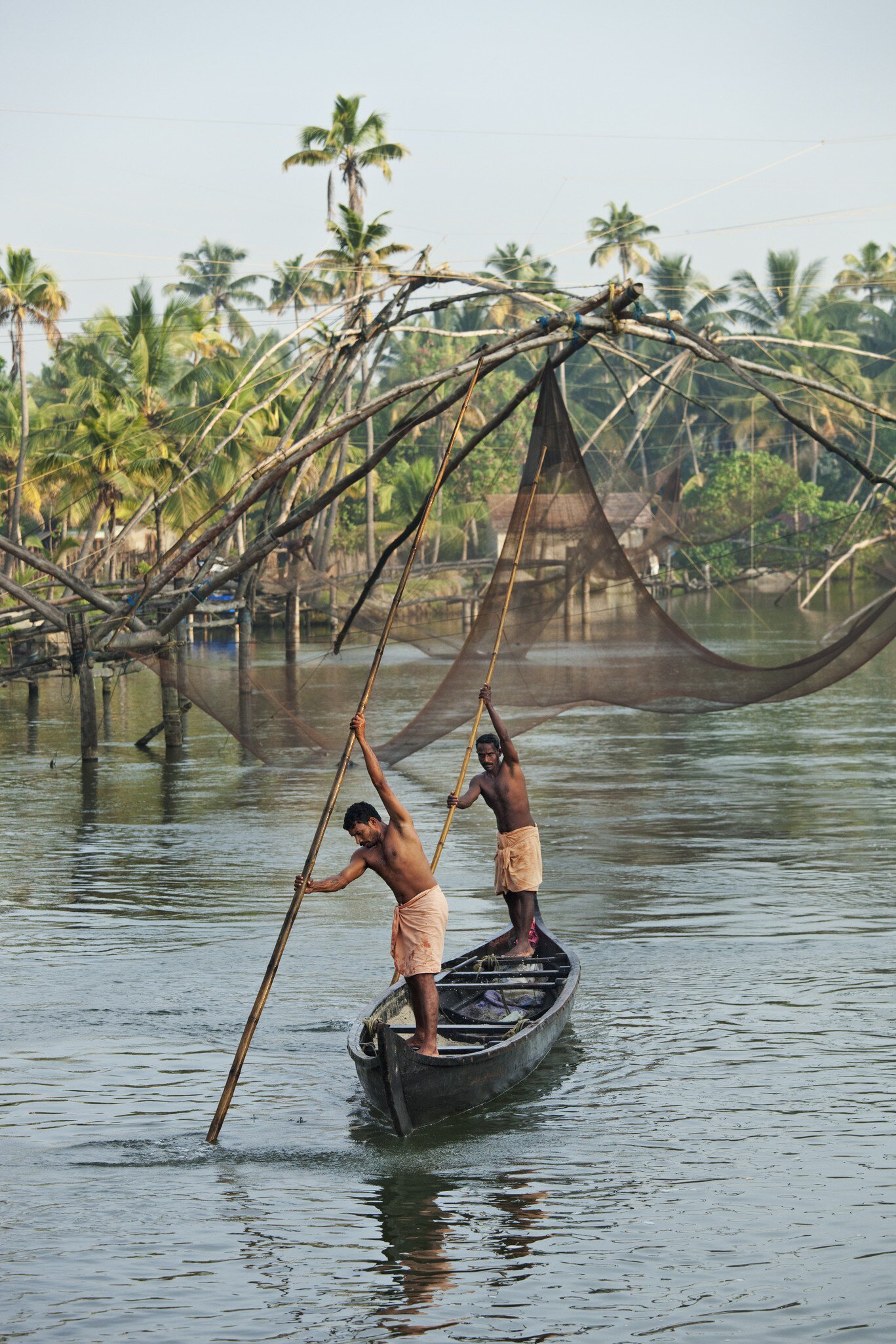 Two bare-chested men in orange lungis push long bamboo poles to steer a canoe past large fishing nets and palm trees.