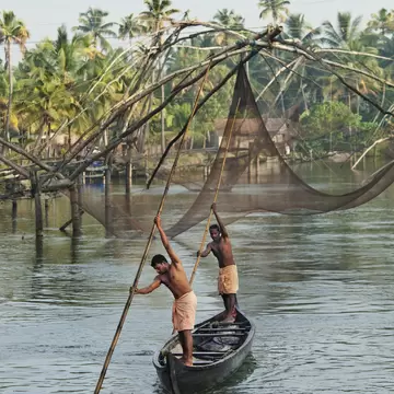 Two bare-chested men in orange lungis push long bamboo poles to steer a canoe past large fishing nets and palm trees.