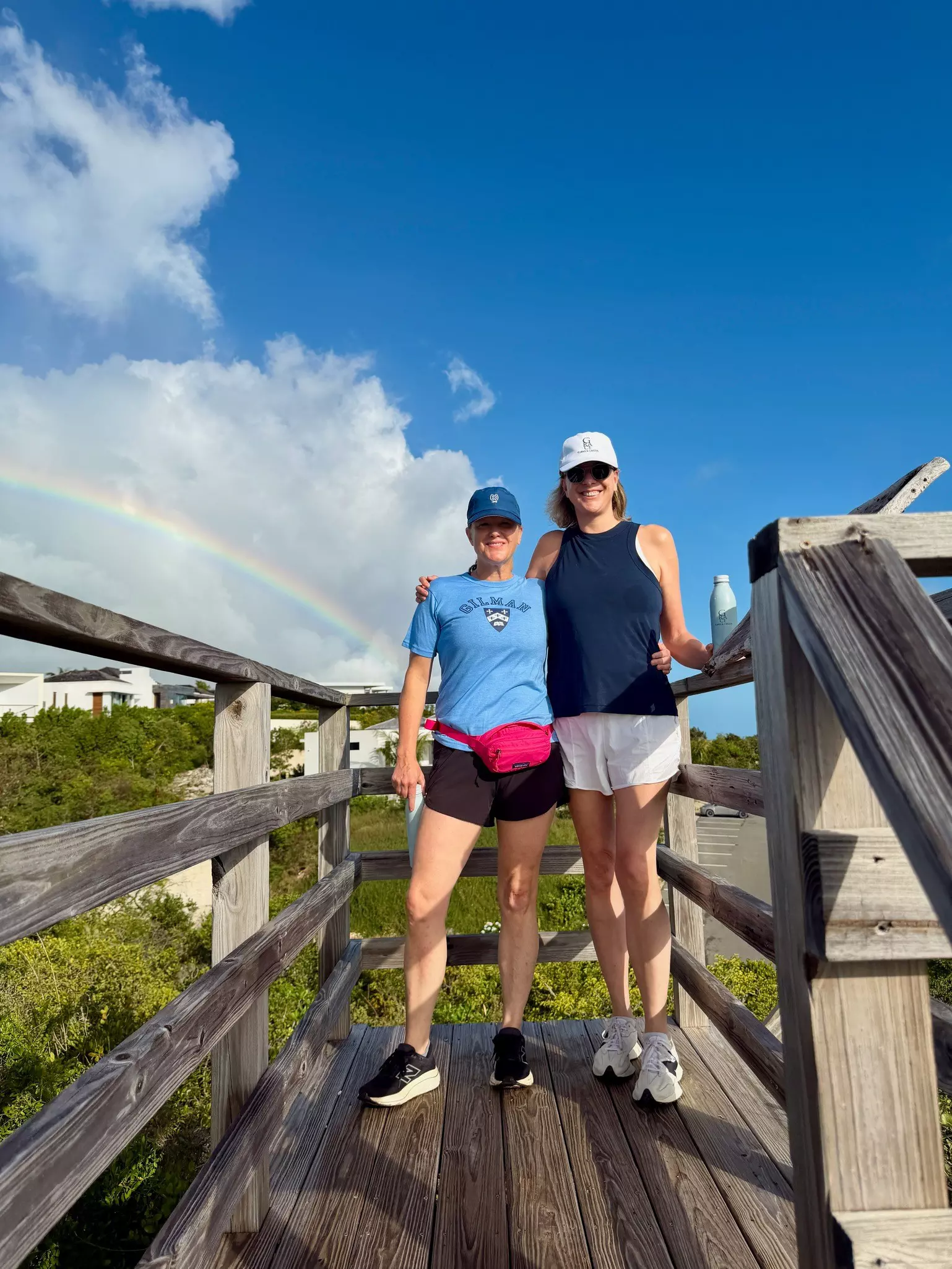 Two women stand together on a wooden boardwalk and smile for the camera. There's a rainbow across the sky behind them