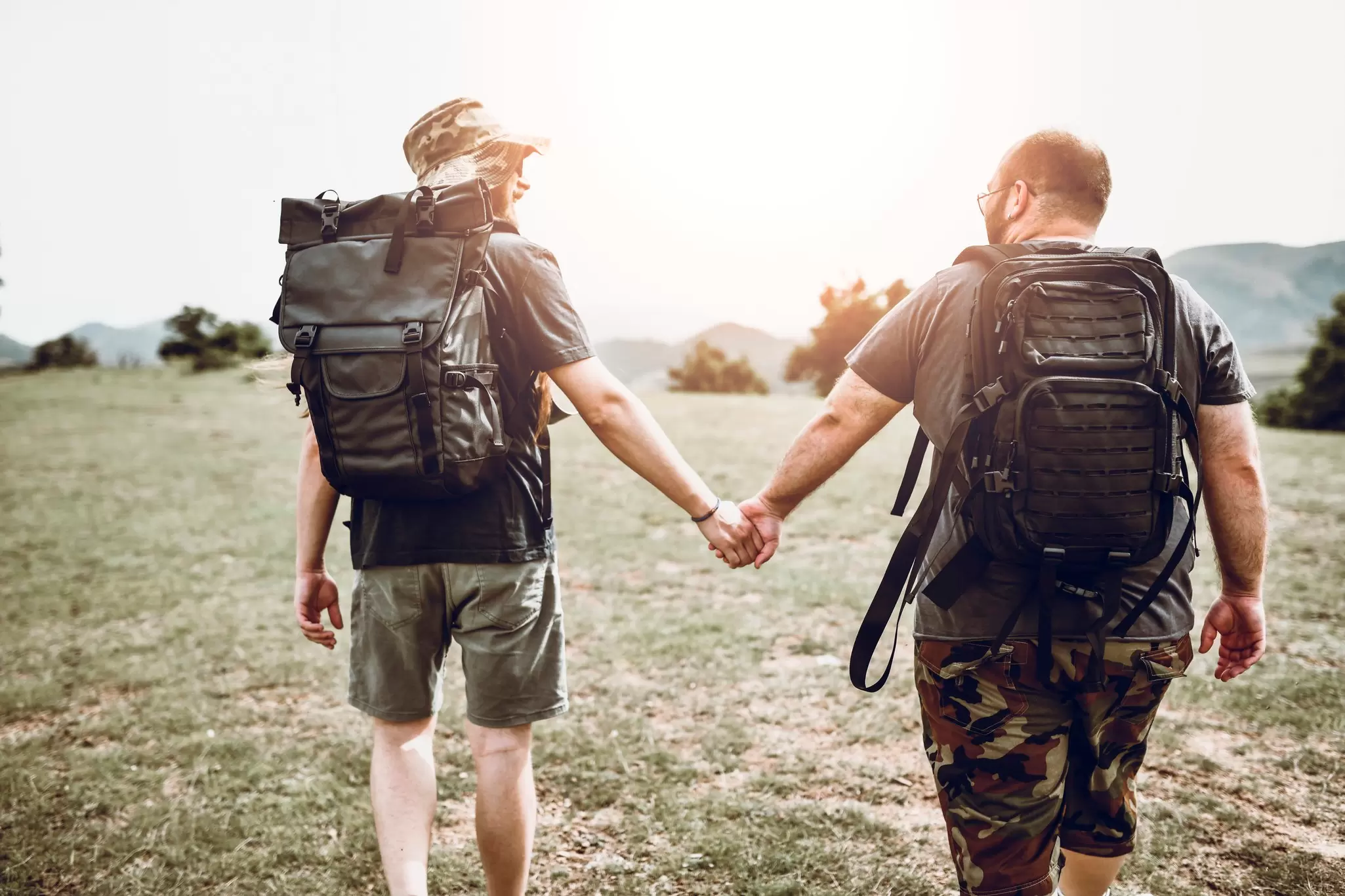 Two hikers with backpacks holding hands from behind as they walk across a grassy hillside at sunset