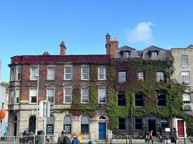 Pedestrians cross a city street in front of buildings covered in ivy.
