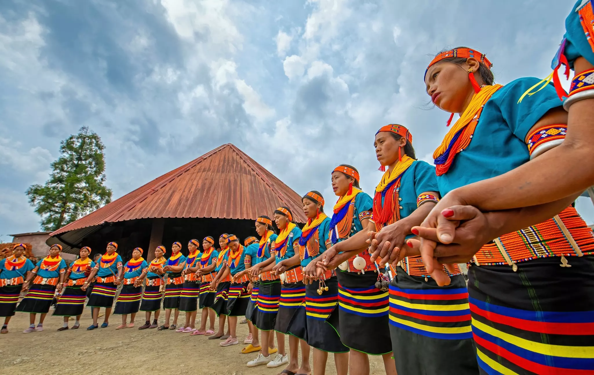 A group of women wearing their traditional tribal dress of colorful stripes and beads stand in a semicircle and perform at a festival.