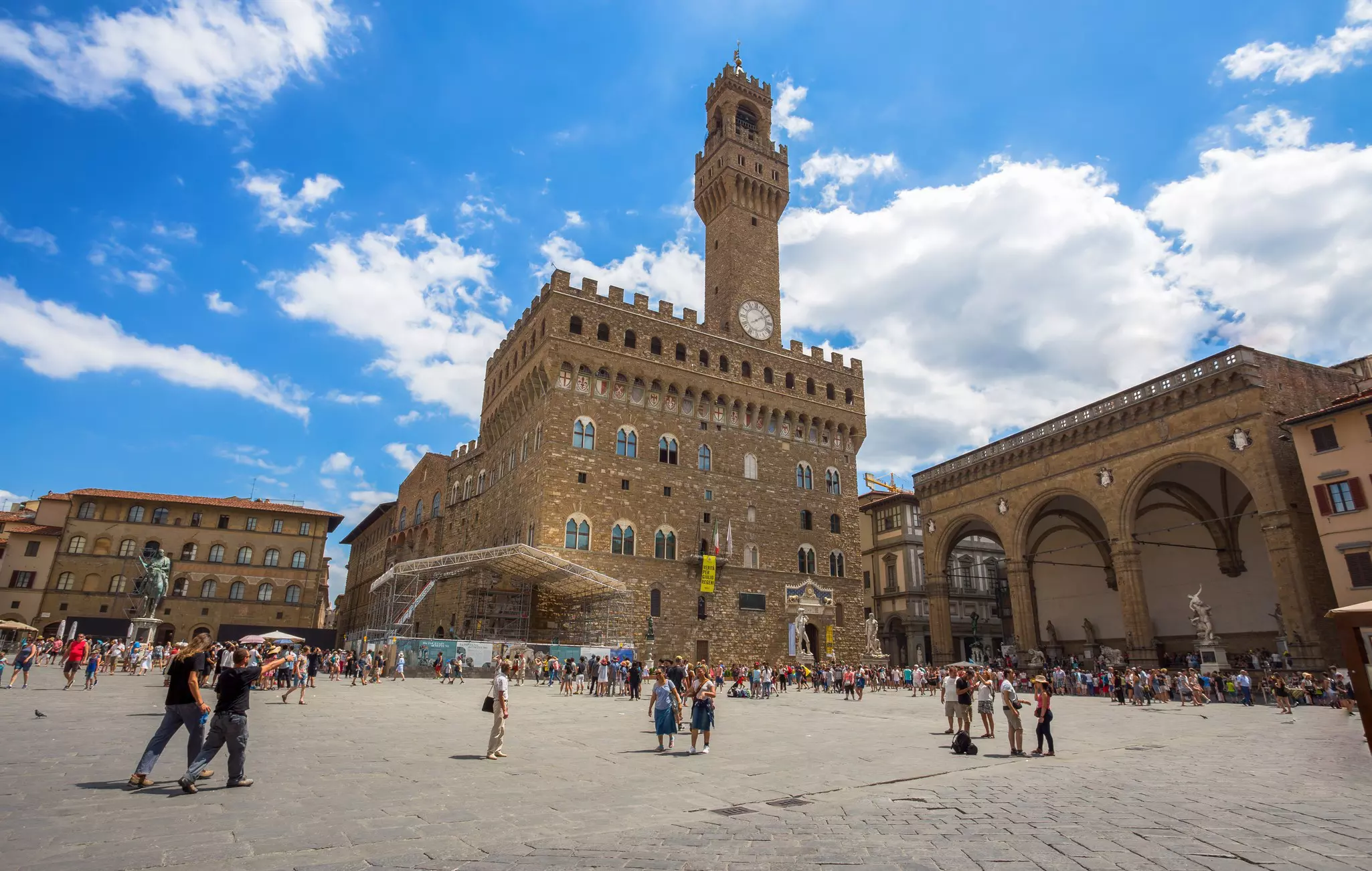 People walking in a plaza in front of large buildings, one with a clock tower
