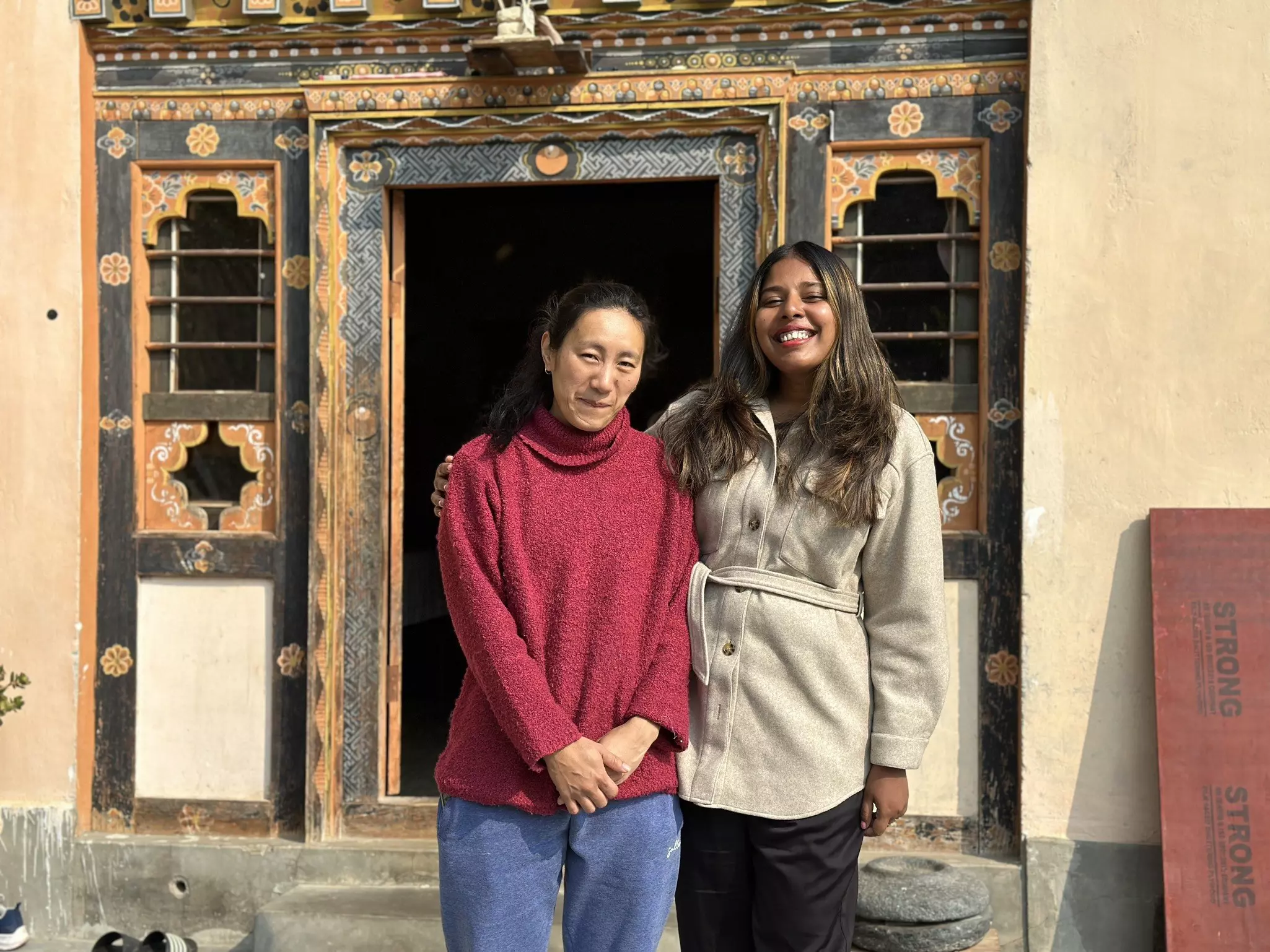 Two women stand together outside a small house with an ornate doorway.