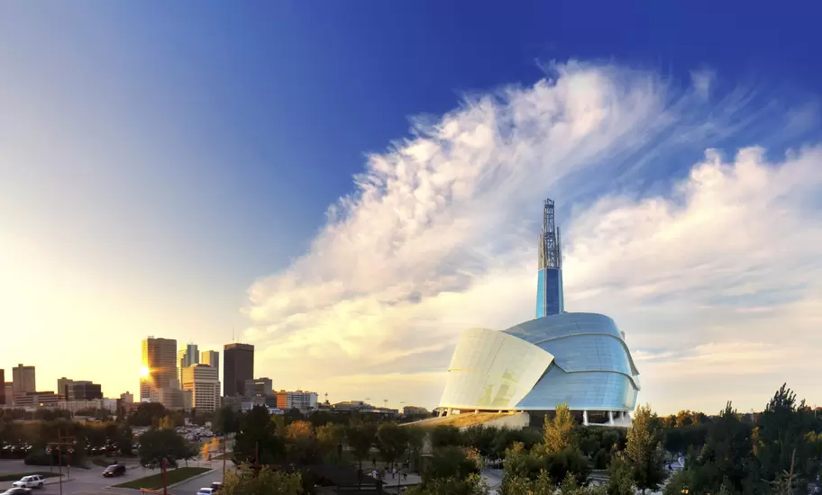 Modern glass-and-stone museum with a tall spire glowing at sunset beside a downtown skyline and wispy clouds