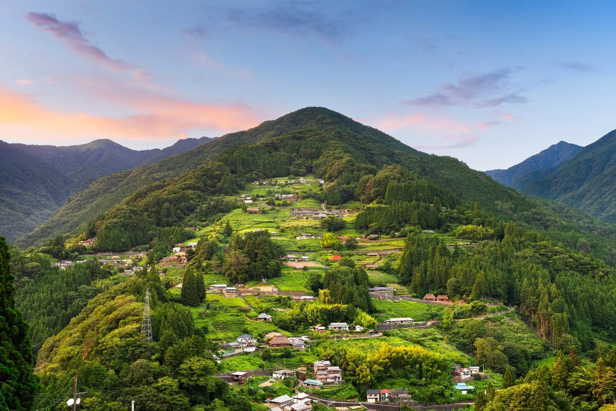 A tree-covered hillside has an area cleared with grass and scattered buildings.