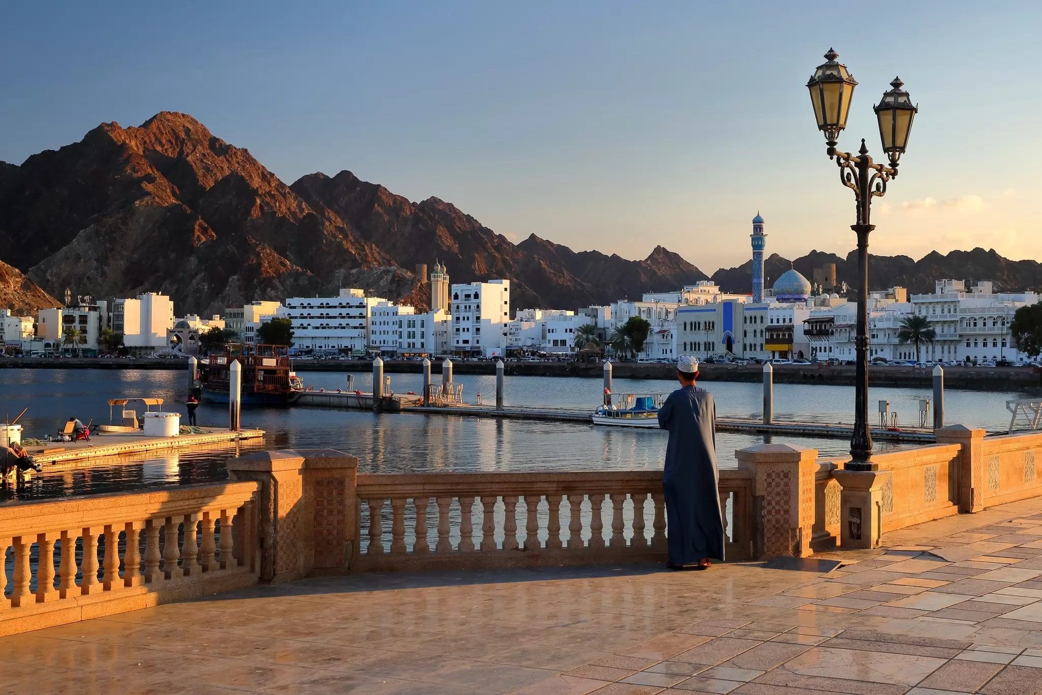 A man stands at a waterfront looking across the harbor towards a mosque and row of white buildings as the sun sets: the corniched, Muscat, Oman